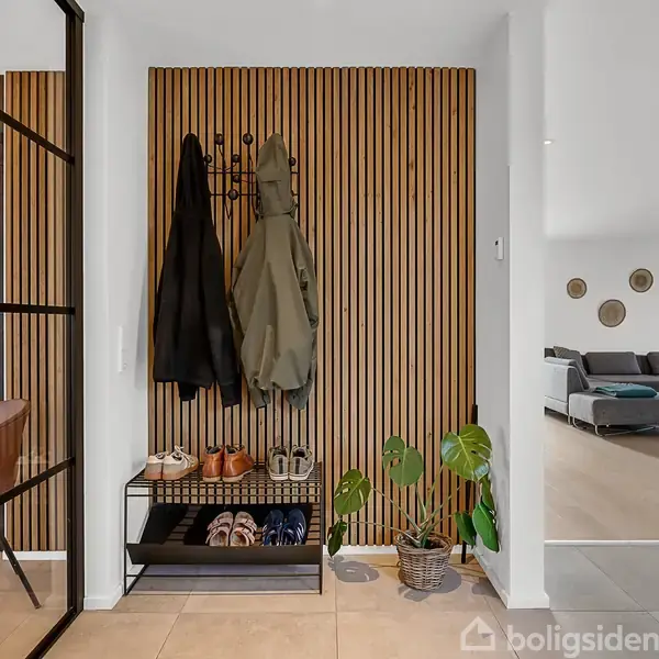 En entré med trækrogpanel og frakker hængende. Skohylde med sko og en plante ved siden af. Til højre ses en stue med grå sofa og vægdekorationer.