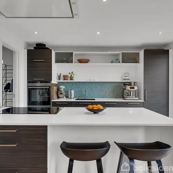 Et moderne køkken med en hvid ø, to sorte stole og en frugtskål. I baggrunden er der en ovn, skabe, en kaffemaskine og et lille vindue.