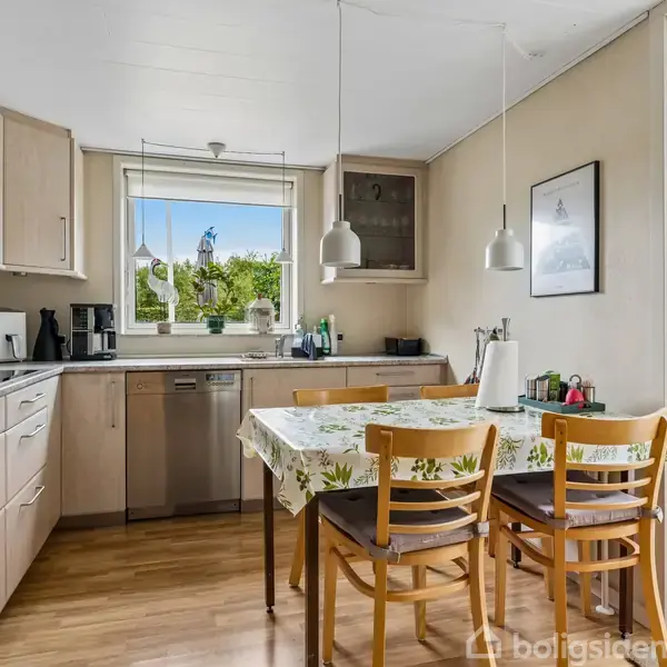 Et moderne køkken med lyse træskabe og stålapparater. Et bord med tre stole står foran et vindue, der viser udsyn til en grøn have. En vase og køkkenredskaber står på bordet.