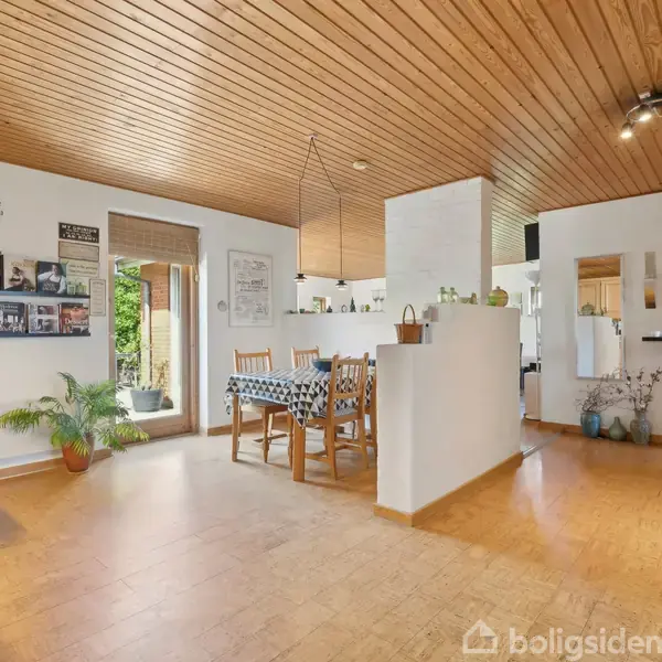 Stue med spisebord og stole, dækket med sort/hvid ternet dug. Omkring er planter, fotos på væggen og en træbeklædt loft. Rummet modtager naturligt lys fra glasdøren.