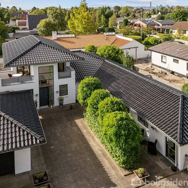 Et stort, moderne hus med sort tag ligger omgivet af frodige træer og velplejede haver i et kystnært boligområde med udsigt over havet i baggrunden.