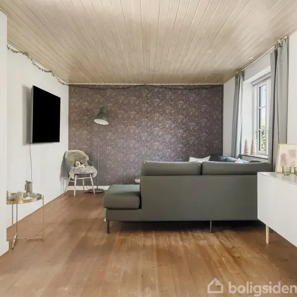 Grå sofa i moderne stue står mod en væg, flankeret af et tv på væggen og en stående lampe. Stuemiljøet har trægulv og store vinduer med naturligt lys.