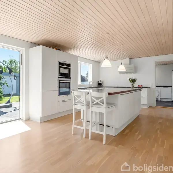 Køkken med hvidt køkkenbord ø, omgivet af høje stole. Store vinduer til venstre åbner til en solrig have. Lyse trægulve og moderne pendellamper i et minimalistisk rum.