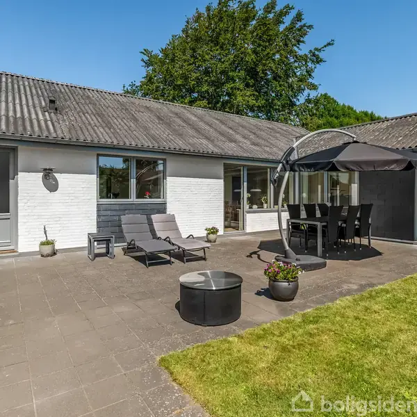Huset med hvid facade har en terrasse med solstole og et spisebord under en parasol. Omgivet af grøn græsplæne, lyse omgivelse og blå himmel ses i baggrunden.