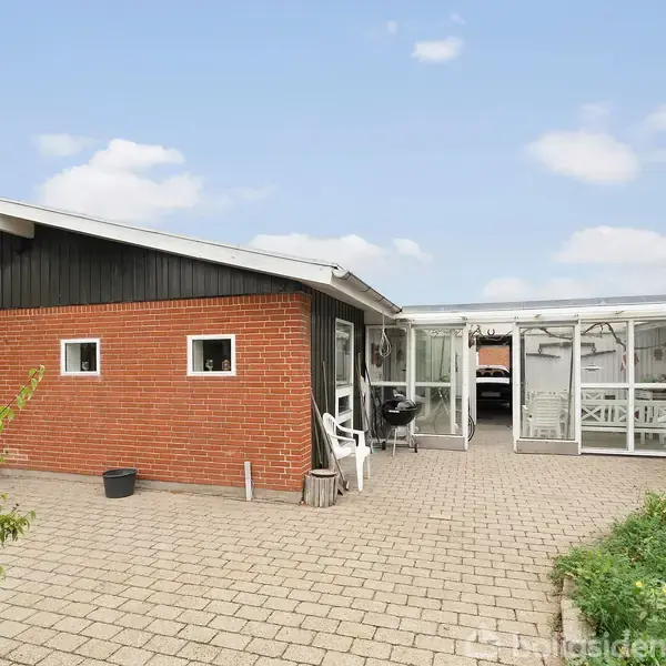 En gårdhave foran et hus med orange murstensvæg og sorte paneler. Flere hvide havestole og bord står nær en glasdørsparti. Gårdspladsen er flisebelagt, omgivet af grønne buske.