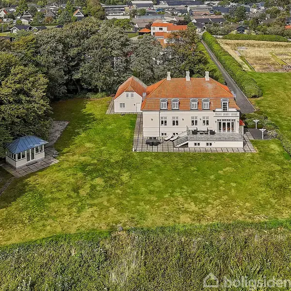 En stor hvid bygning med rødt tag står på en grøn plæne. Til venstre ses en lysthus, og i baggrunden er der en skov og flere bygninger i det fjerne.