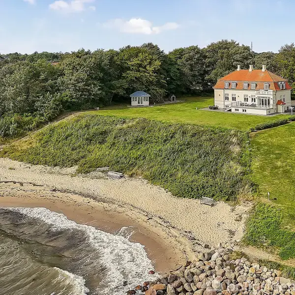 Stor villa med rødtag ligger fredeligt på bakketop. Den omkranses af frodige græsplæner og skovområder, mens havet og en sandstrand ligger nedenfor med bølger skvulpende mod kysten.