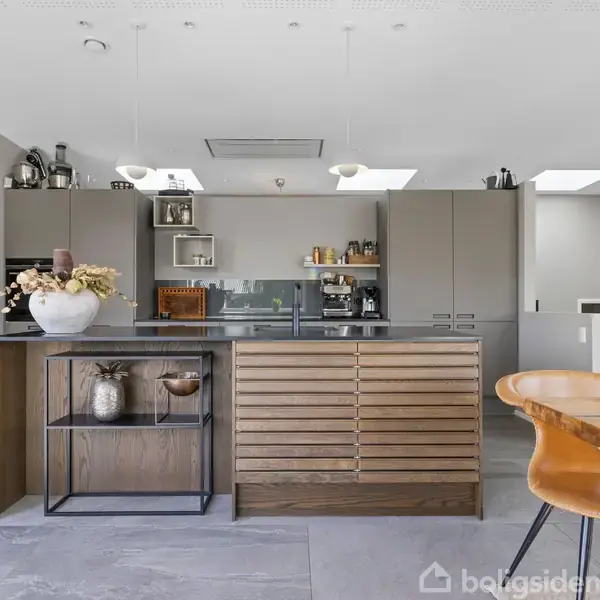 Et moderne køkken med en central køkkenø i trælook. Omkranset af grå skabe, vinopbevaring og spiseplads med brune stole. Lysekroner hænger fra loftet.