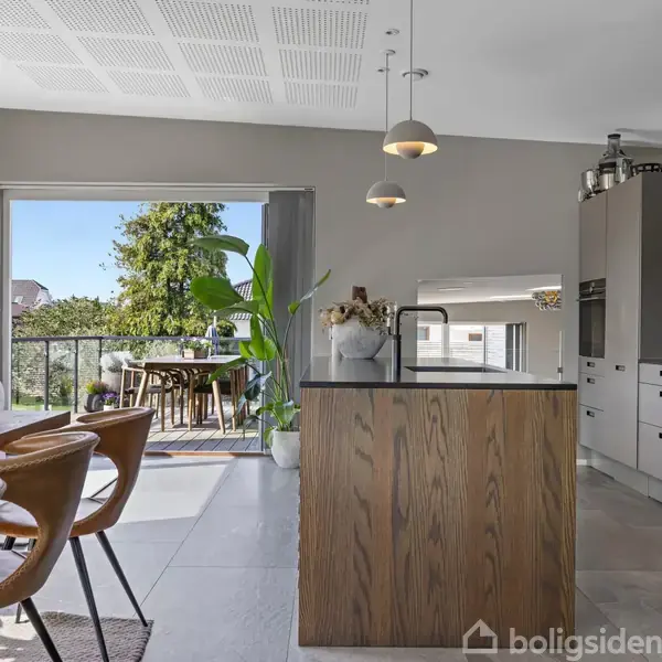 Et moderne køkken med ø, omgivet af spisebord og stole, åbner ud til en terrasse med udsigt over træer. Lyse farver og minimalistisk design præger rummet.