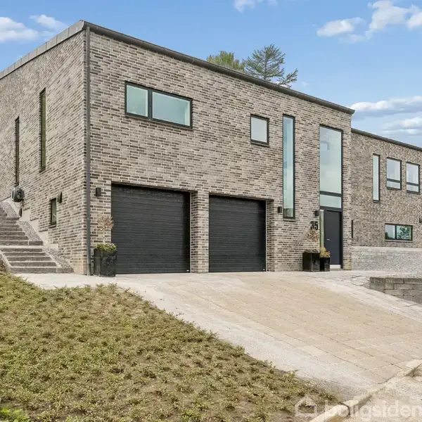 Et moderne hus lavet af grå mursten står på en skrånende indkørsel. Huset har dobbelte garageporte og store vinduer. Omgivet af grønne buske og træer.