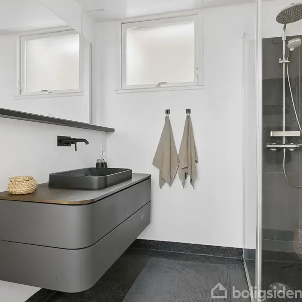 En carport med fladt tag og nummeret "24" er delvist overdækket. Den er omgivet af flisebelagt indkørsel og røde murstensbygninger i et boligområde med træer i baggrunden.