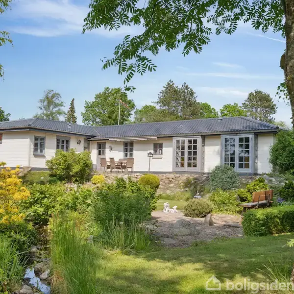 Et hvidt hus med sorte tagsten står i en frodig have, omgivet af træer og buske. Foran huset er der en terrasse og en dekorativ have med sten og græsplæne.