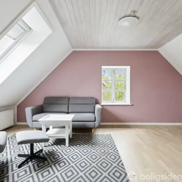 Grå sofa står i et lyserødt værelse med skråvægge. Foran er et hvidt sofabord og et mønstret tæppe. Hvidt loft med loftslampe og lille vindue i baggrunden.