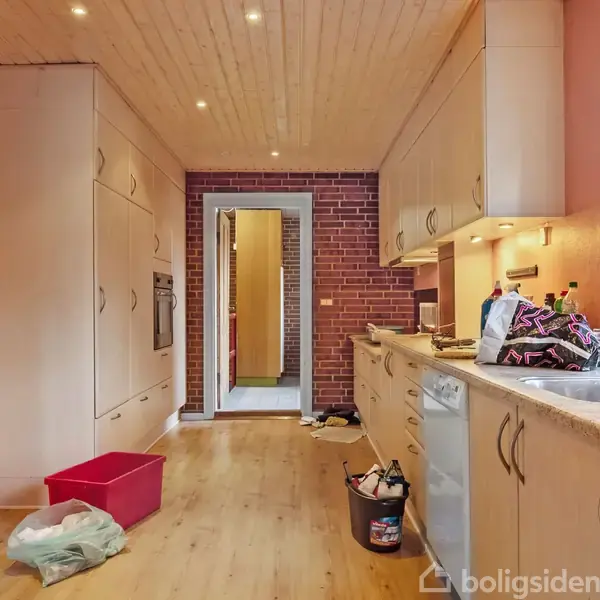 Køkken med lyst trægulv og loft, hvide skabe, og vask på højre side. Rød murstensvæg omkring døråbning for enden. Rodede genstande på bordpladen, spand på gulvet.