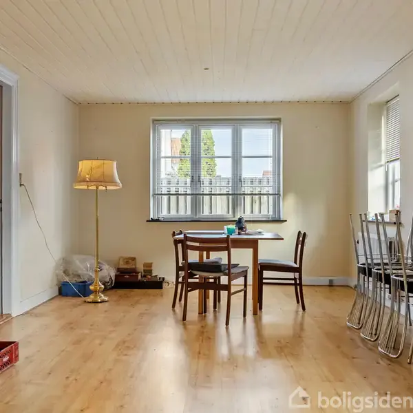 Et rundt bord med stole står i et rum med trægulv. En gulvlampe til venstre er tændt, og der er stablede stole til højre ved vinduerne.