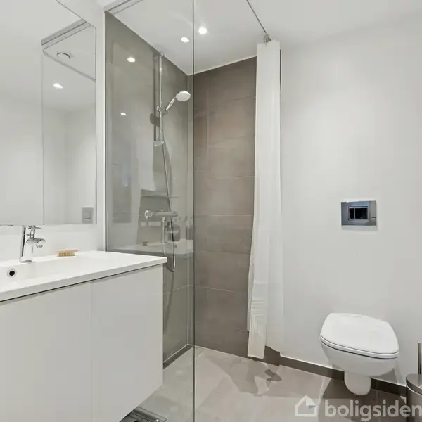 Badeværelse med vask og skab integreret i en spejlbeklædt væg. Brusekabine med glasvæg og bruser. Toilet monteret på en lys, ren væg. Minimalistisk og moderne indretning.