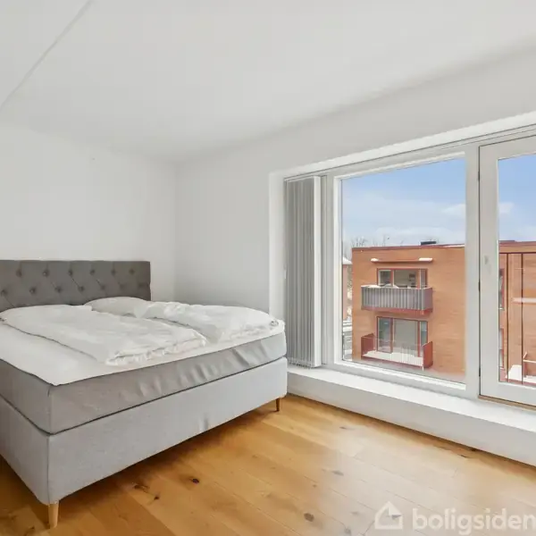 En dobbeltseng med gråt hovedgærde står i et lyst værelse med trægulv. Store vinduer viser en udsigt til et rødt murstensbyggeri udenfor.