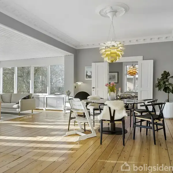 Spisebord med sorte stole og hvidt betræk står i solrig stue med træloft, store vinduer og lyse træplanker. Et moderne lysarmatur hænger over bordet, omgivet af planter.