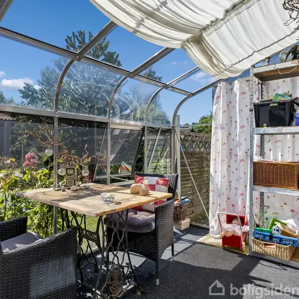 Glasoverdækket udestue med to kurvestole omkring et bord, omgivet af blomster i en have. Hylde med bøger og kurve. Lys gardin hænger fra loftet.