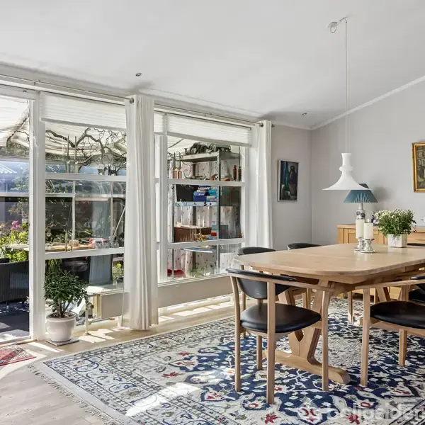 Spisebord med fire stole står på et orientalsk mønstret tæppe. Store vinduer og glasdøre åbner mod en terrasse med planter. Lysekrone hænger over bordet, moderne og lyst design.