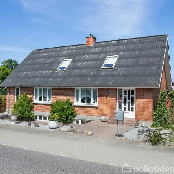 Rødt murstenshus med sort tag har hvide vinduer og en skorsten, omringet af en velplejet have. Det ligger ved en stille vej under en blå himmel.