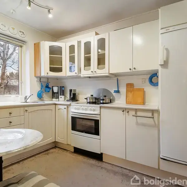 Et lyst køkken med hvide skabe og en lille spiseplads. En ovn står midt i rummet med gryder ovenpå. Et stort køleskab står til højre ved siden af køkkenbordet.