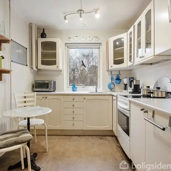 Et hvidt køkken med et lille rundt bord og to stole. Skabene er åbne med glaslåger, og der er en ovn på højre side. I baggrunden ses et vindue med udsigt til træer. Hylder på væggen har dekorative genstande.