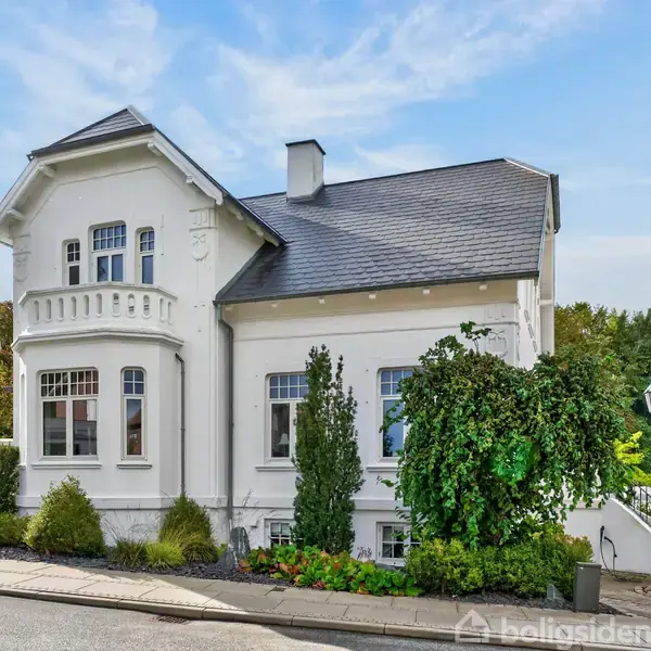 Hvid villa står stille ved en vej, omgivet af velplejede buske og træer. Detaljeret vinduesparti ses på facade, med klar blå himmel i baggrunden.