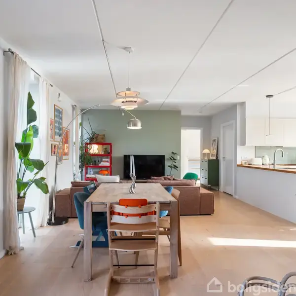 Spisebord med stole står i et lyst, moderne køkken-alrum. Stuen har en sofa foran et TV. Grønne planter og kunst pryder væggene, mens store vinduer lader lys ind.