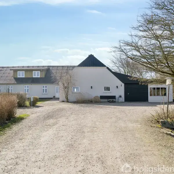 Et stort, hvidt hus med sort tag står for enden af en bred grusvej. Omkring huset er der træer og buske, der skaber en landlig atmosfære.