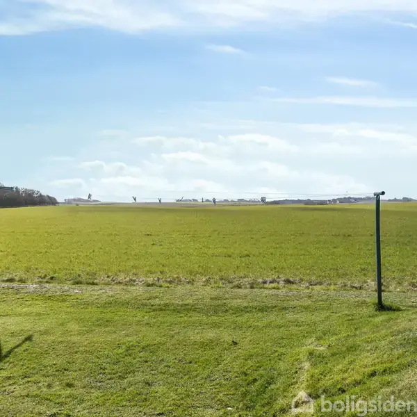 Grønt markeret område strækker sig ud under en klar blå himmel. To sorte lygtepæle står uden aktivitet langs markens kant. Udkig til flere bygninger i det fjerne.