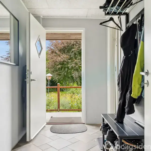 En entré med åbne døre, viser en udsigt til en frodig have. Jakker hænger fra en knagerække, og en bænk med opbevaring står ved siden af.