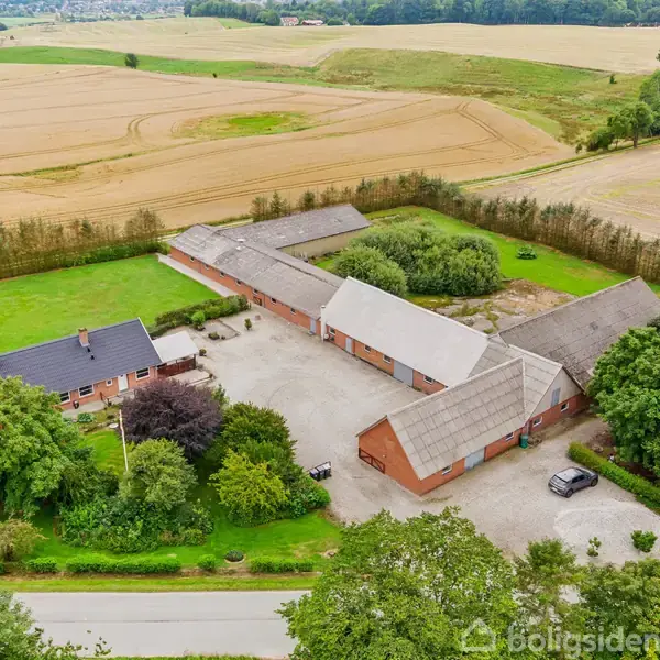 Gårde ligger i landligt område omgivet af marker. Flere bygninger danner en gårdsplads. Hus omgivet af træer til venstre, og parkeret bil ved siden af indkørsel.