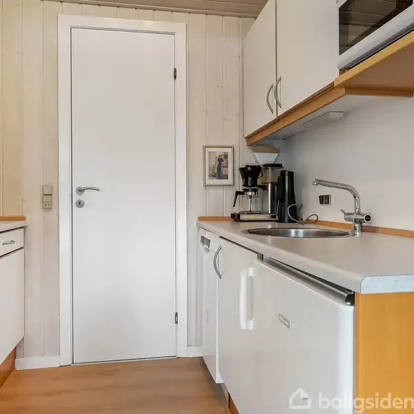 Et køkken med hvide skabe og træbordplader. Der står en kaffemaskine ved vasken. Ovnen og kogepladen er til venstre. En lukket dør ses i baggrunden.