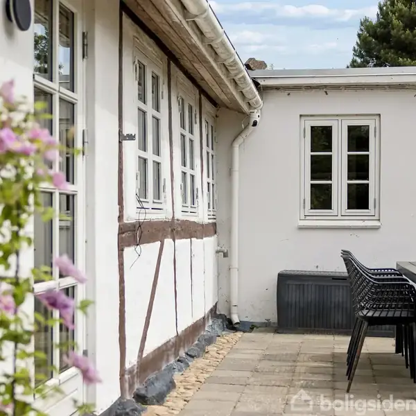 Hus med bindingsværk, stilrene hvide vægge og sprossede vinduer. Et havebord med stole står på en flisebelagt terrasse. En blomstrende plante er til venstre i billedet.