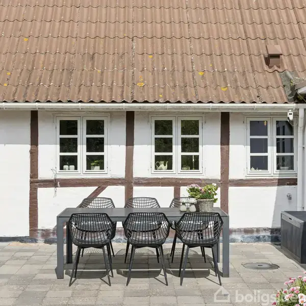 Et udendørs spisebord med seks stole står på en terrasse foran en bindingsværksbygning med hvide vægge og sprossede vinduer. Omkring bordet er blomsterpotter og flisebelægning.