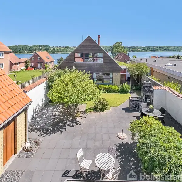 Et hyggeligt gårdmiljø med havemøbler, placeret på en flisebelagt terrasse. Omkring ses velplejede buske og træer. I baggrunden står et brunt, trekantet hus med røde tage og en udsigt over vandet.