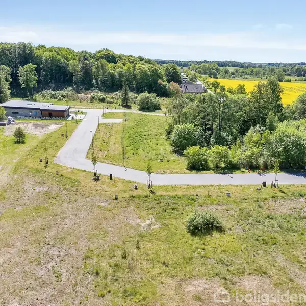 En bygning ligger ved en asfaltvej i et åbent landskab med grønne marker og skov i baggrunden. Du kan se marker med gule blomster i det fjerne.