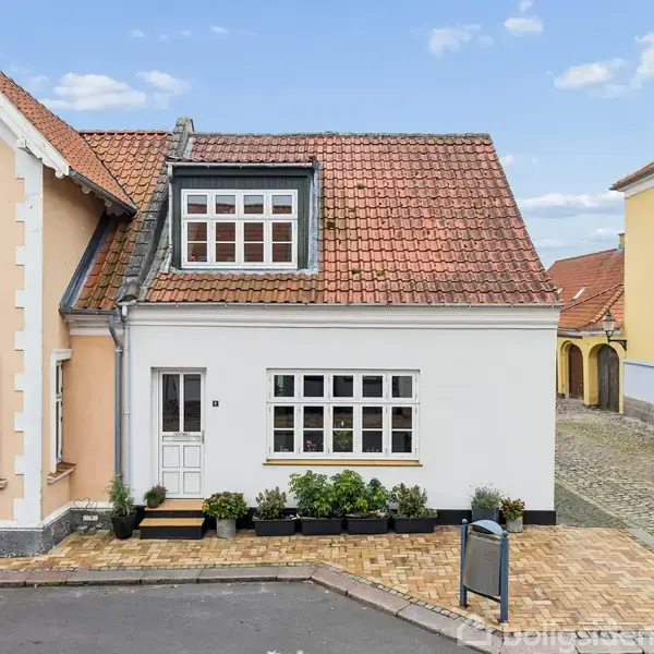 Hvidt hus med rødt tegltag, store vinduer og en lille trappe med blomsterkrukker foran indgangen. Står på en brostensbelagt gade ved siden af et lyserødt hus og en gadelampe.