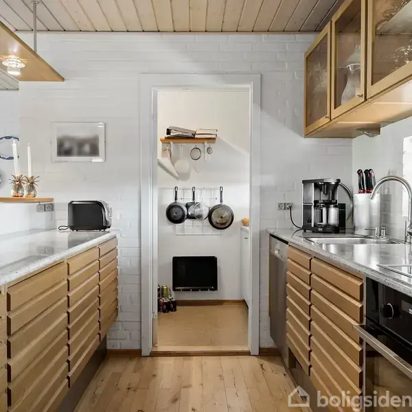 Køkken med træskabe og hvidt loft, byder på moderne apparater som kaffemaskine og brødrister. Gryder hænger på væggen. Et vindue til højre viser udsigt til grønne træer.