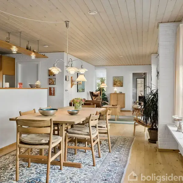 Et spisebord med træstole står på et orientalsk tæppe i en lys stue. Rummet har vinduer med planter, træloft og åbner mod et afslapningsområde med sofaer.