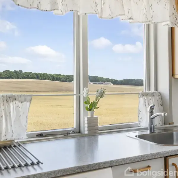 Køkkenvask med hvid bordplade og stålvask ved vindue. Blomster i vase. Udsigt over gul kornmark med træer i horisonten under blå himmel med skyer.