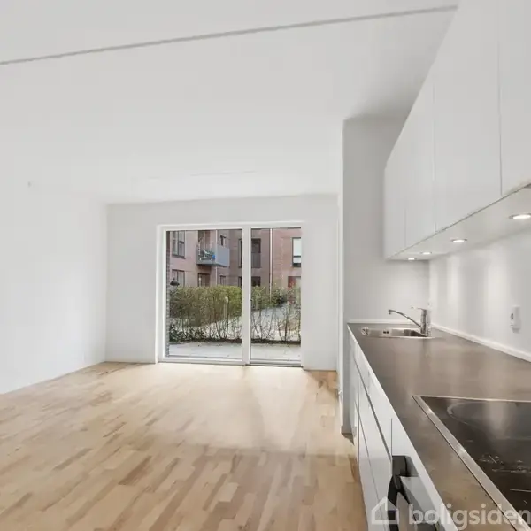 En moderne, lys lejlighedsstue er forbundet med et åbent køkken. Store vinduer åbner mod en balkon med udsigt til en bygning og en hæk. Gulvet er af lyst træ.
