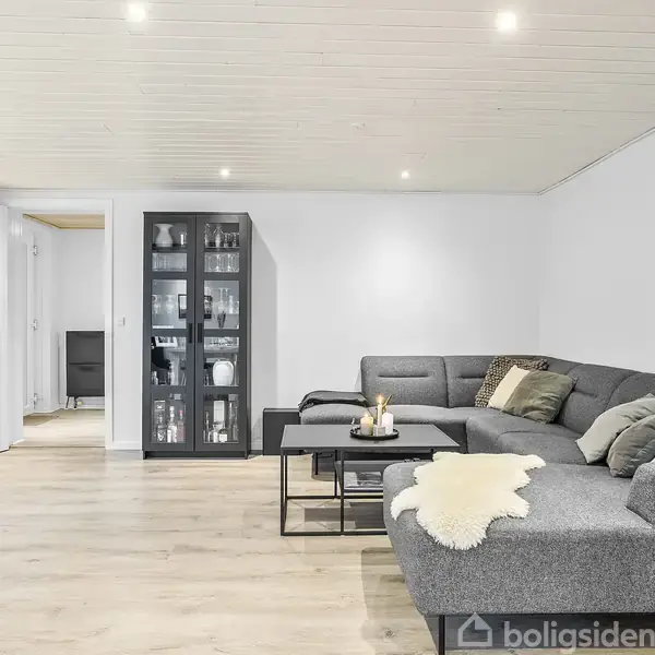 En grå, L-formet sofa med puder står i en lys stue. Foran den er et sofabord med lys og dekoration. En glasvitrine placeret ved væggen med pyntegenstande.