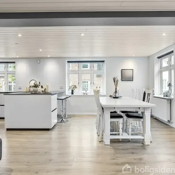 Stort, lyst køkken-alrum med hvidt køkkenbord og køkkenø. Spisebord med stole placeret i hjørnet ved vinduer. Trægulv og hvide vægge skaber en moderne, luftig atmosfære.