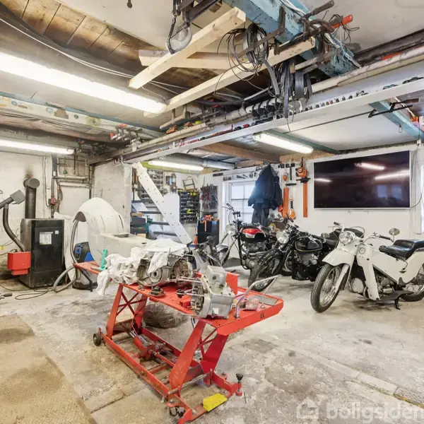 Motorcykler står parkeret i et værksted med redskaber og maskiner hængende fra loftet. I midten er et arbejdsbord med motordele. Væggene har hylder og værktøj.