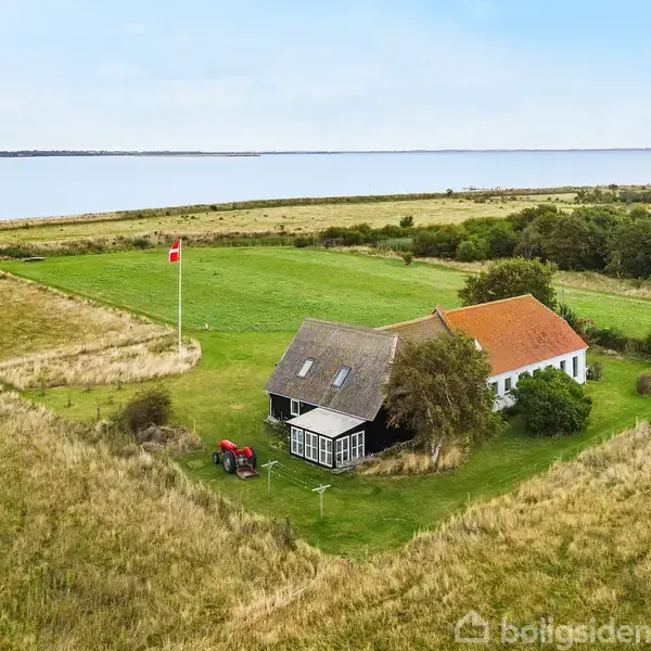 Et hus med rød og gråtag, beliggende på en grøn mark ved en sø. En flagstang med dansk flag og en rød traktor står ved siden af.