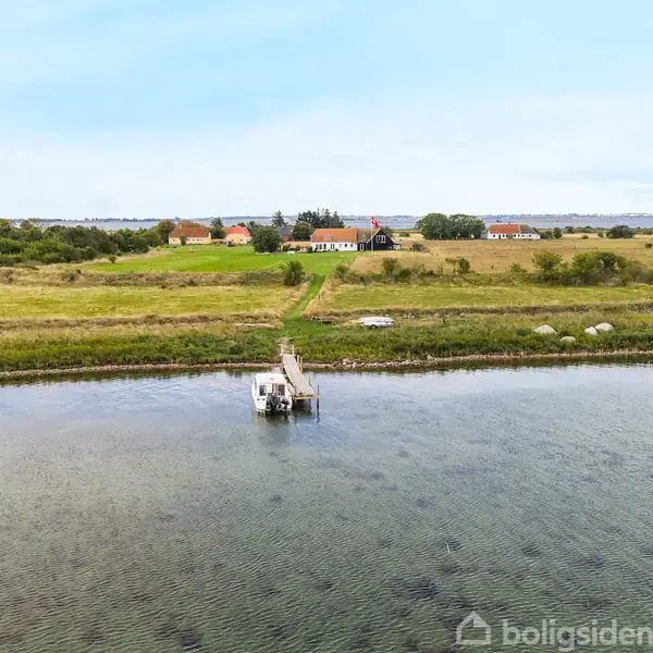 En lille båd er fortøjet til en træbro langs en kyst. Broen fører til en grøn mark, hvor der i baggrunden er nogle huse og bevoksning.