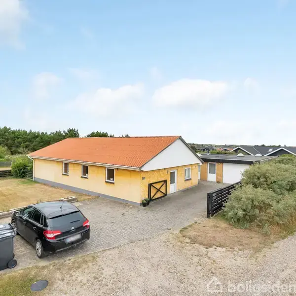 Gul murstenshus med rødt tag står på en grund med flisebelagt indkørsel. En sort bil er parkeret foran huset. Omgivelserne inkluderer buske, græs og andre huse i baggrunden.