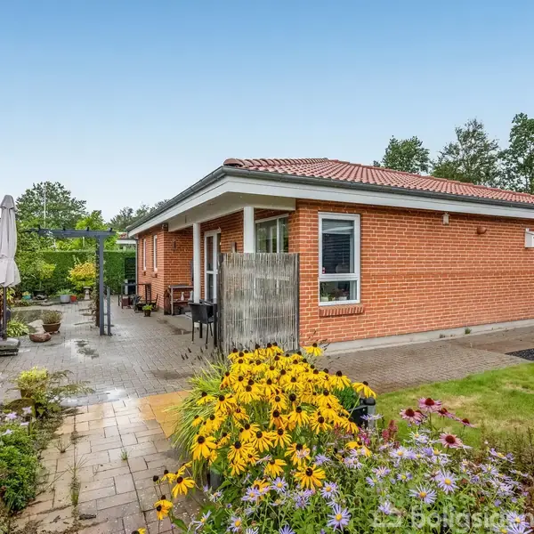 Rødt murstenshus står stille i frodig have. Foran huset blomstrer farverige planter, og en flisesti fører til en terrasse med parasol. Træer omkranser området.